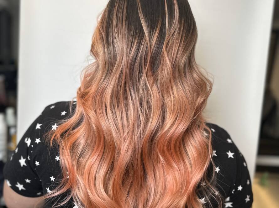 A woman with long flowing hair in a rose gold ombre, transitioning from deep red at the roots to vibrant pink and finally into gold at the ends.