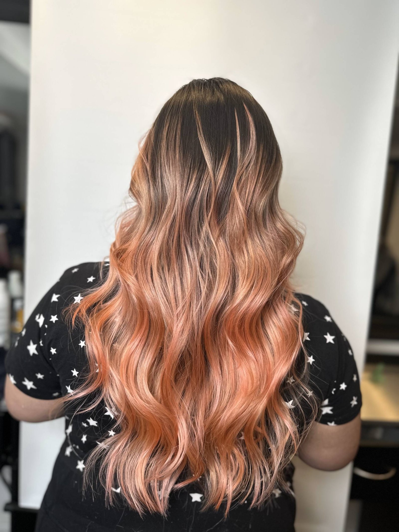 A woman with long flowing hair in a rose gold hair ombre, transitioning from deep red at the roots to vibrant pink and finally into gold at the ends.